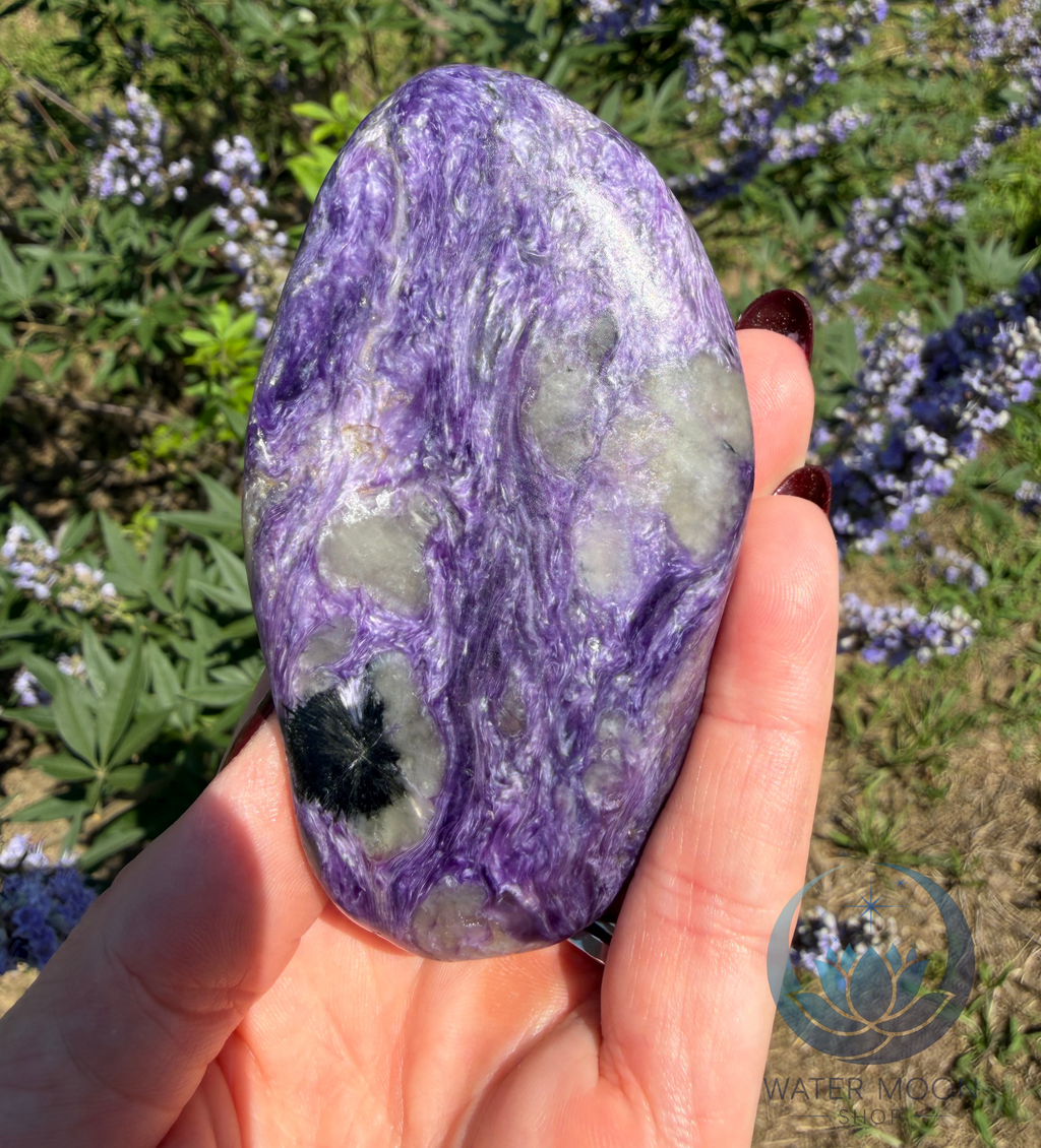 Hand holding a marbled purple and green stone with a natural background of greenery and flowers.