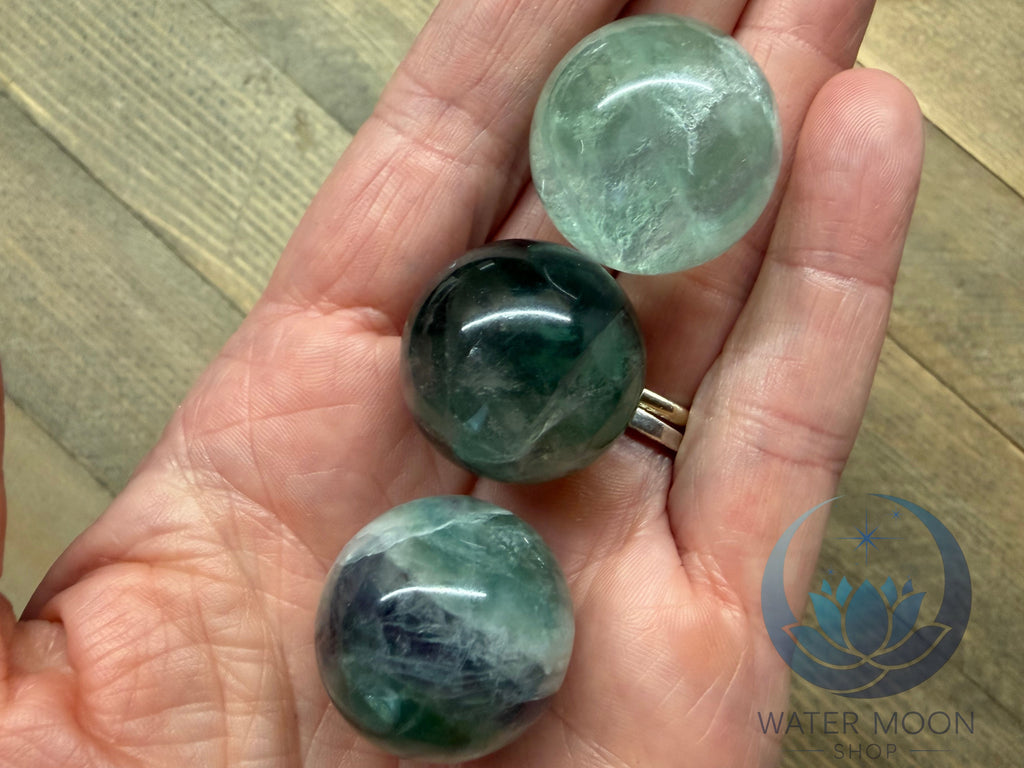 Three green gemstones held in a hand on a wooden surface, with 'Water Moon' logo in the corner.