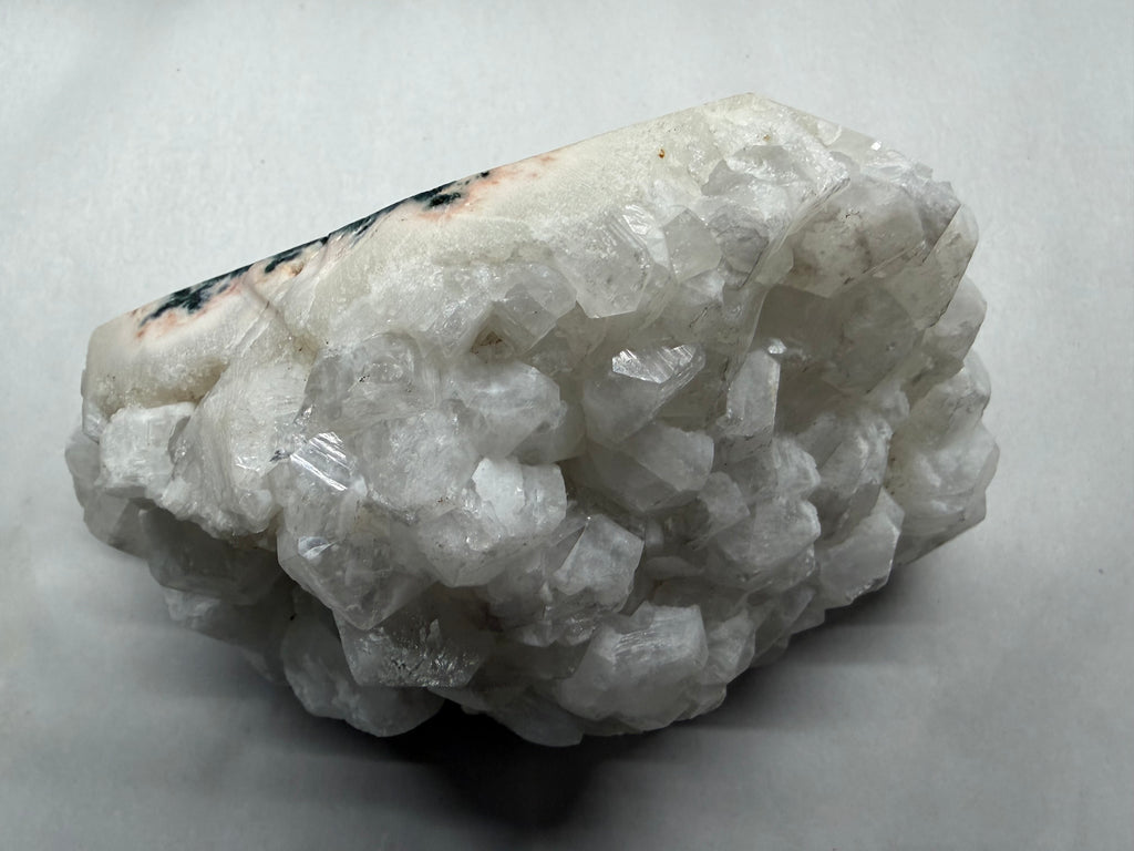A raw mineral crystal specimen featuring dense clusters of clear, faceted white quartz-like crystals on an off-white matrix, with a small dark brown to black area near the top left, photographed on a neutral light gray background.
