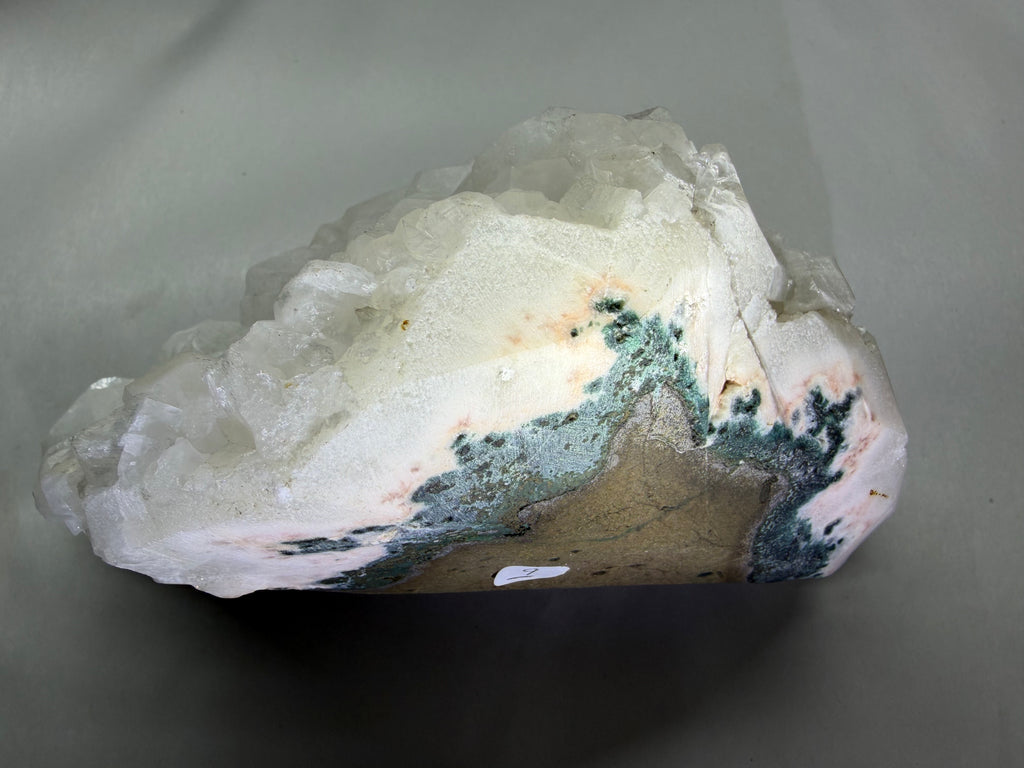 A raw crystal cluster specimen, primarily composed of translucent white quartz, featuring prominent dark green and gray mineral inclusions and a beige matrix, photographed on a neutral gray background.