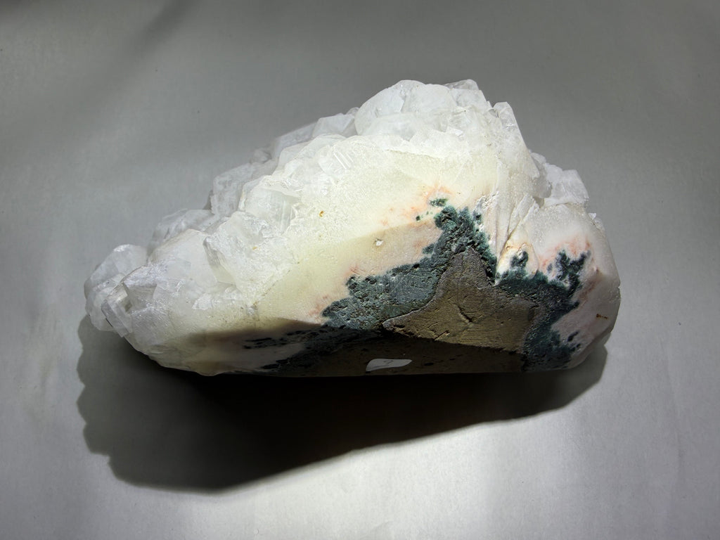 A raw mineral specimen featuring a large cluster of translucent white quartz crystals over a matrix with patches of gray, black, and light pink, resting on a gray surface.