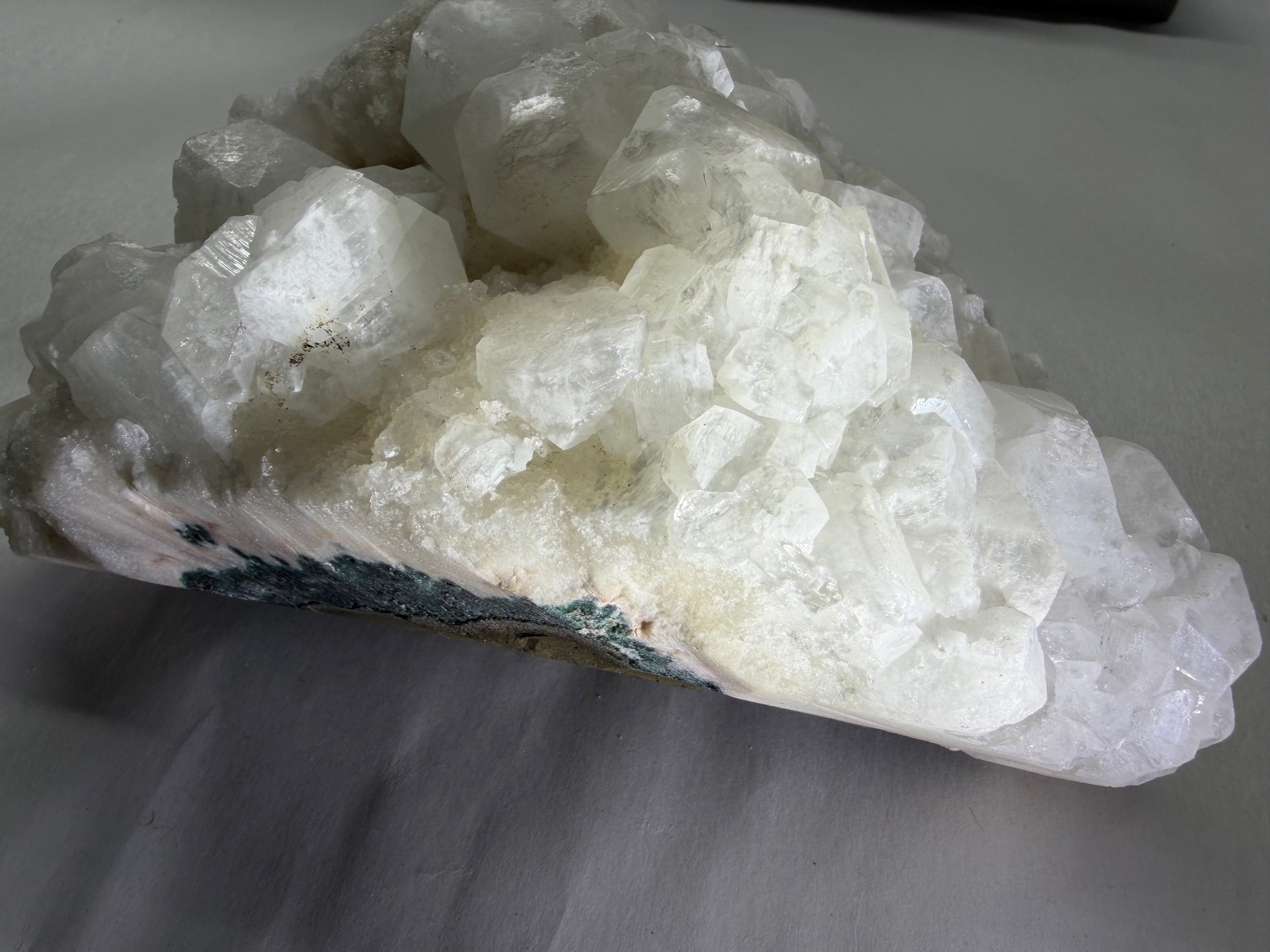 A raw crystal cluster specimen, primarily composed of translucent white quartz-like crystals with a dark green and brown matrix visible along the lower left edge, resting on a gray surface.