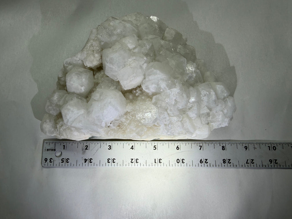 A cluster of rough white crystals or mineral pieces on a light background, with a metal ruler placed underneath to show scale.