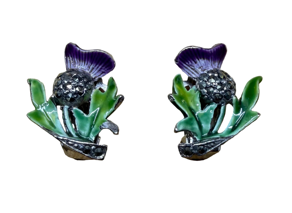 A pair of vintage-style floral enamel pins shaped like purple thistles with green leaves, displayed on a light wood grain background.