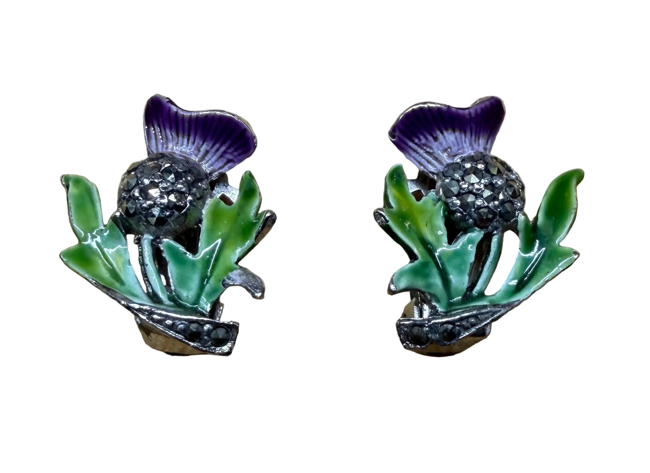 A pair of vintage-style floral enamel pins shaped like purple thistles with green leaves, displayed on a light wood grain background.