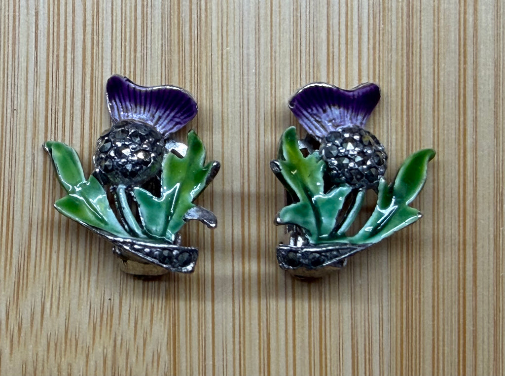 A pair of vintage-style thistle-shaped earrings with purple enamel petals, green leaves, and a dark rhinestone center, displayed on a light wood grain background.