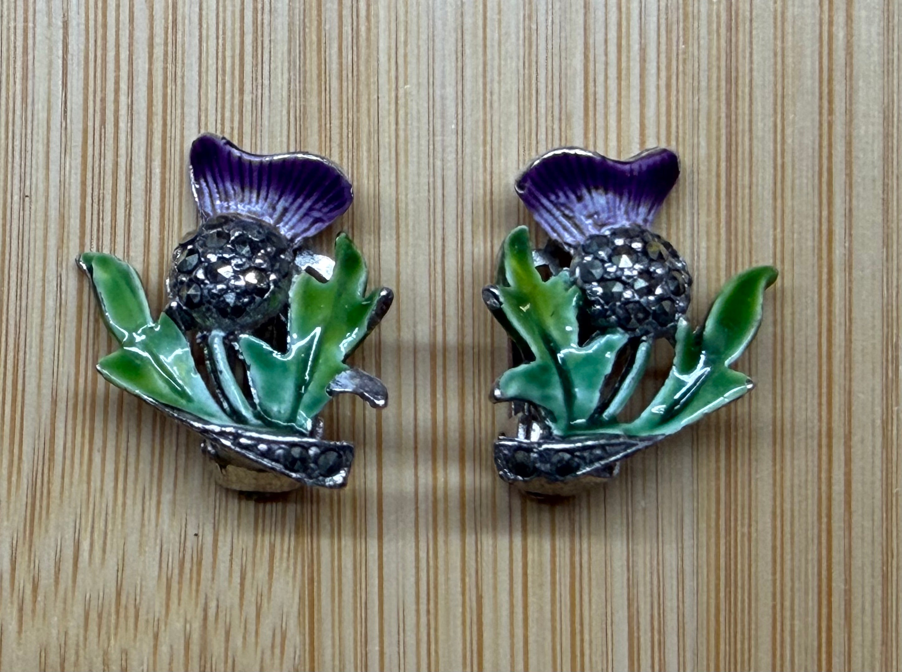 A pair of vintage-style thistle-shaped earrings with purple enamel petals, green leaves, and a dark rhinestone center, displayed on a light wood grain background.