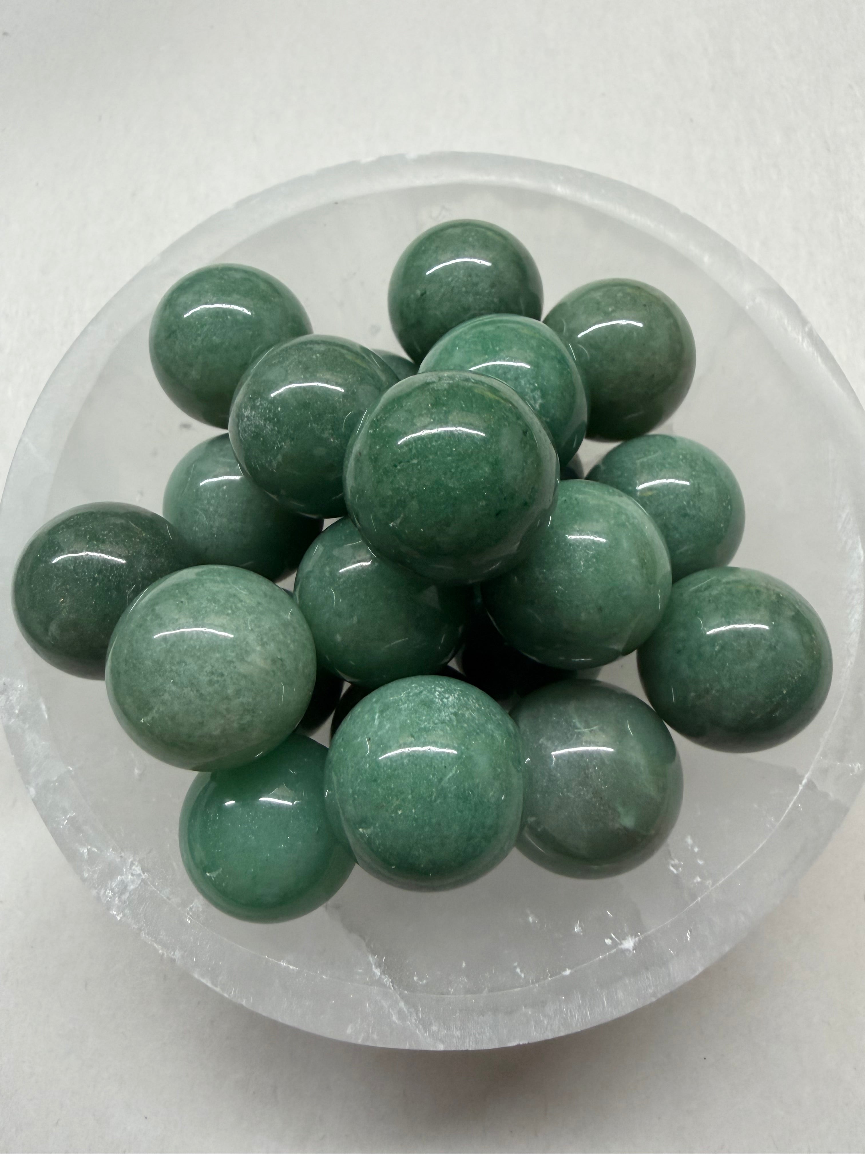 A pile of polished green jade-like round beads displayed inside a clear glass bowl on a light gray background.