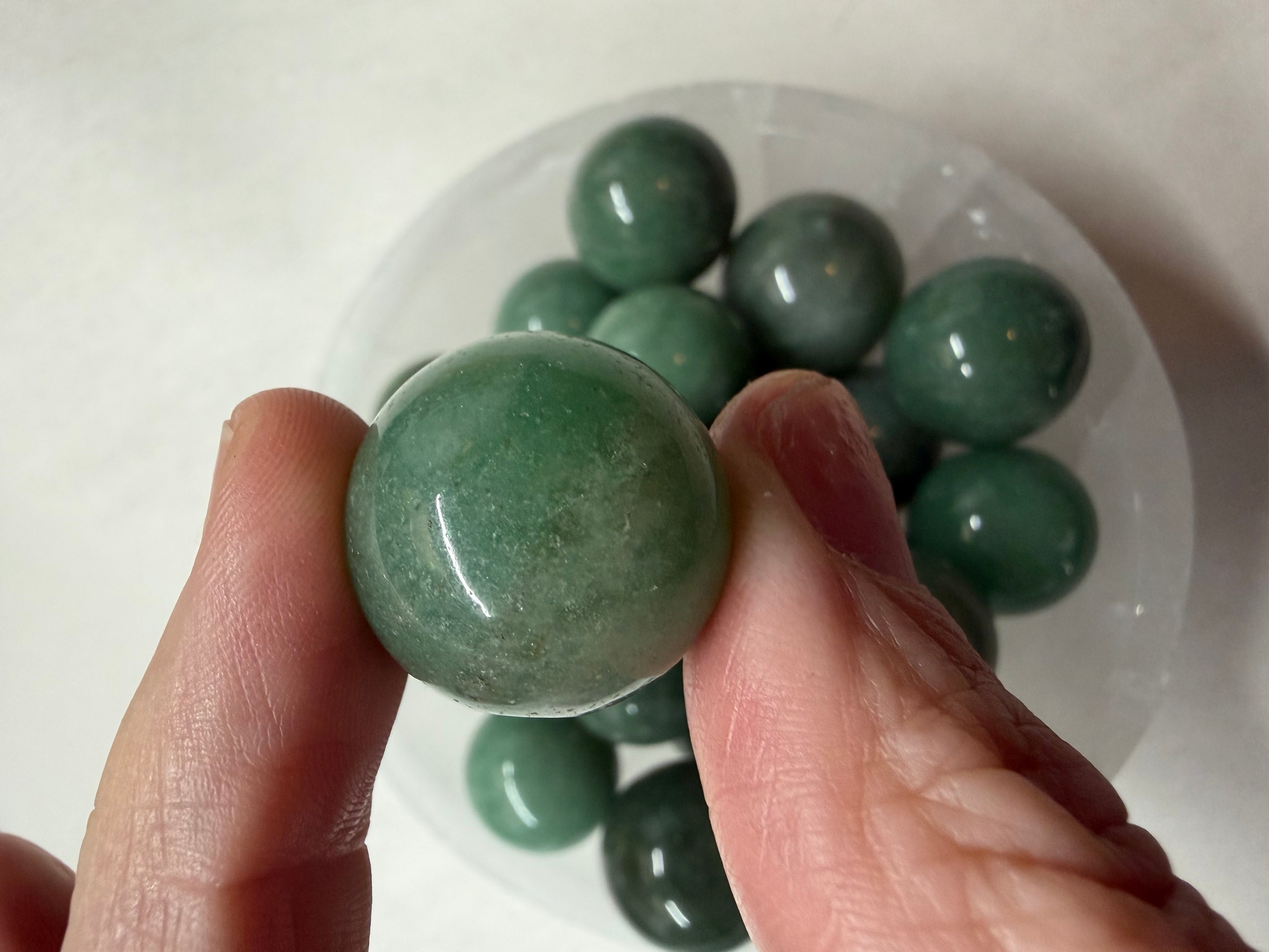 A hand holds a polished green stone sphere in front of a clear plastic container filled with similar green stone balls on a light background.