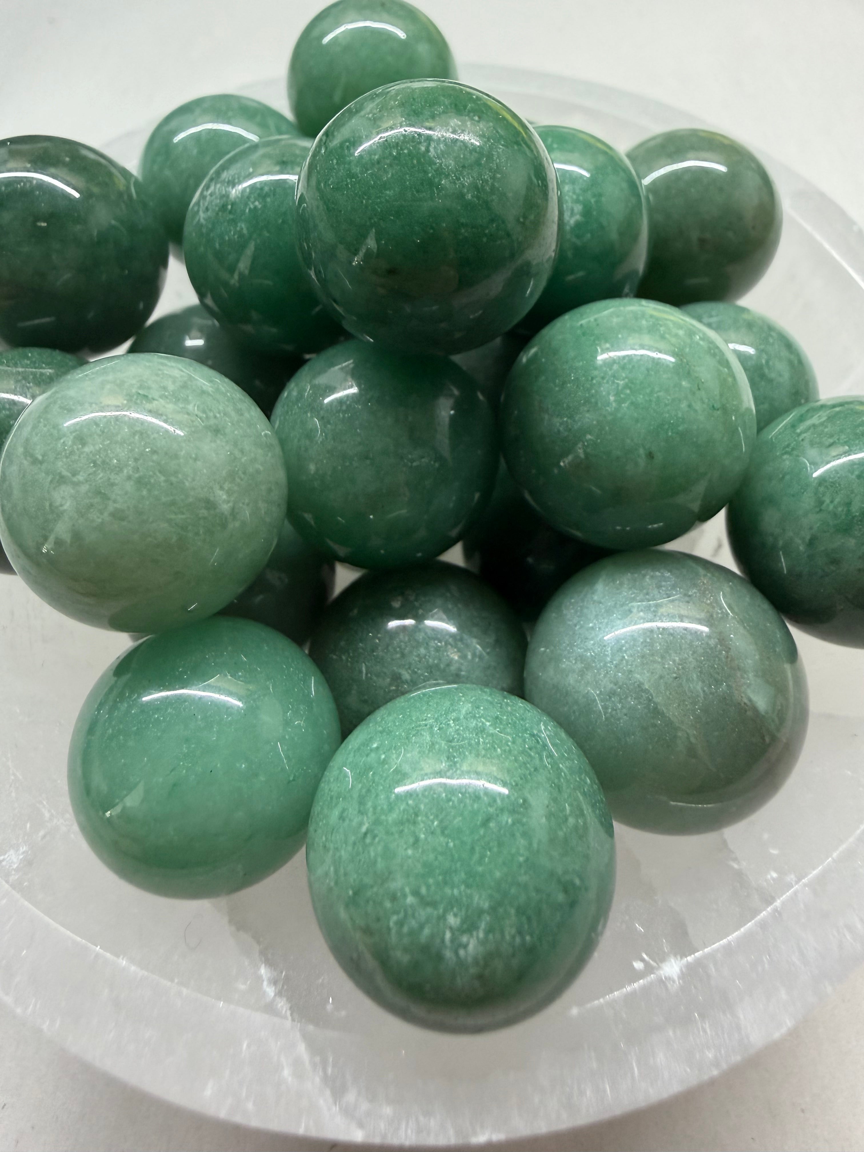 A pile of polished green jade-like round beads or pendants arranged in a clear glass dish on a white surface.