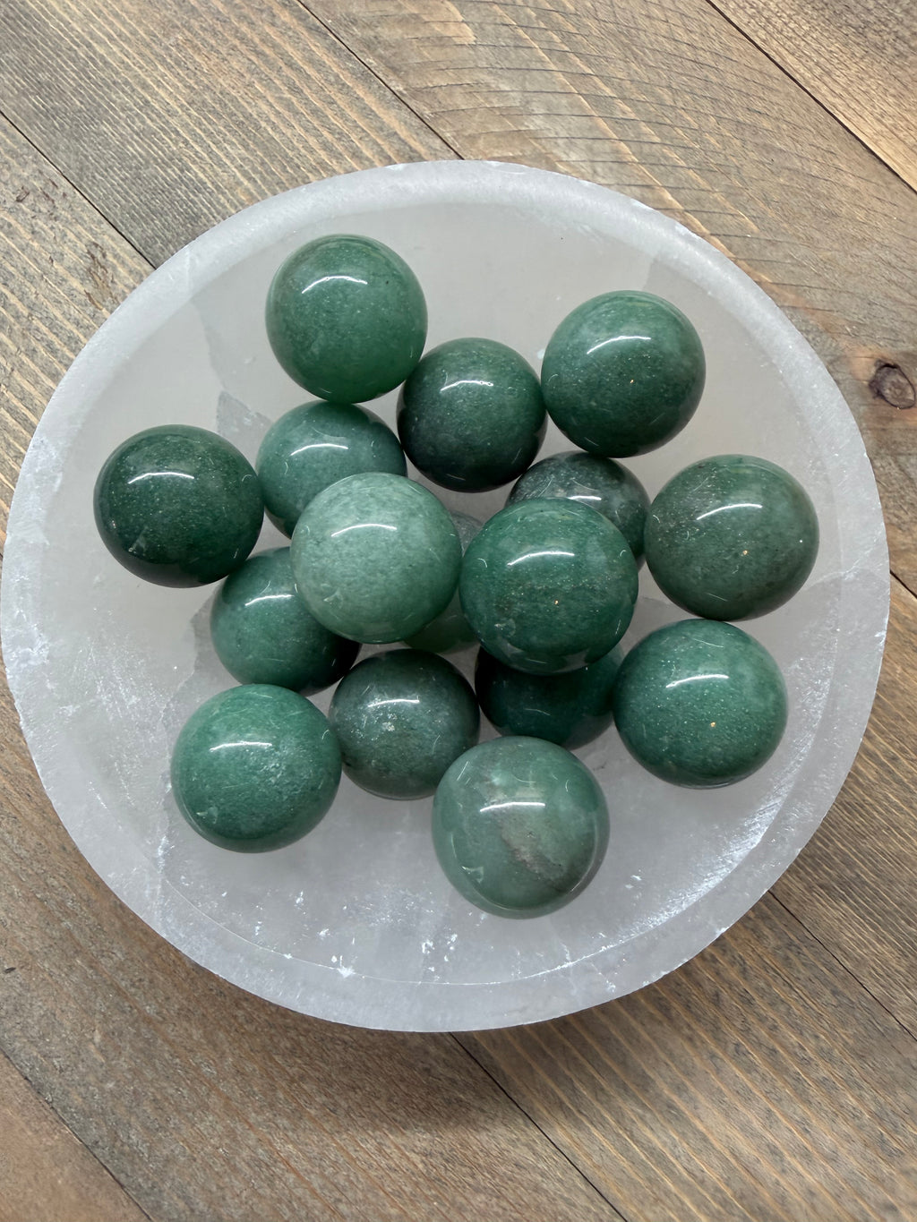 A group of polished dark green crystal balls arranged inside a clear, round crystal bowl on a rustic wooden surface.