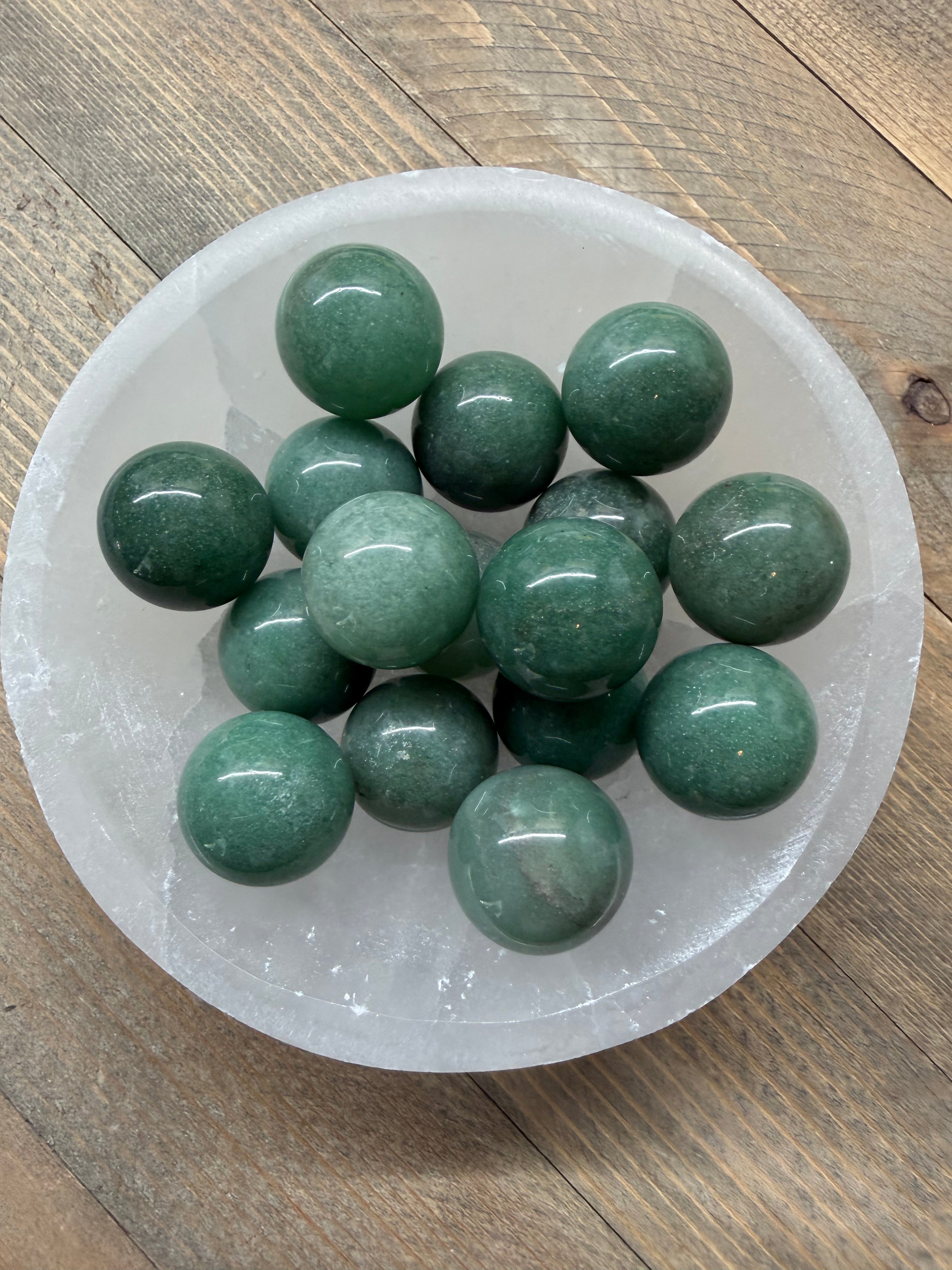 A group of polished dark green crystal balls arranged inside a clear, round crystal bowl on a rustic wooden surface.