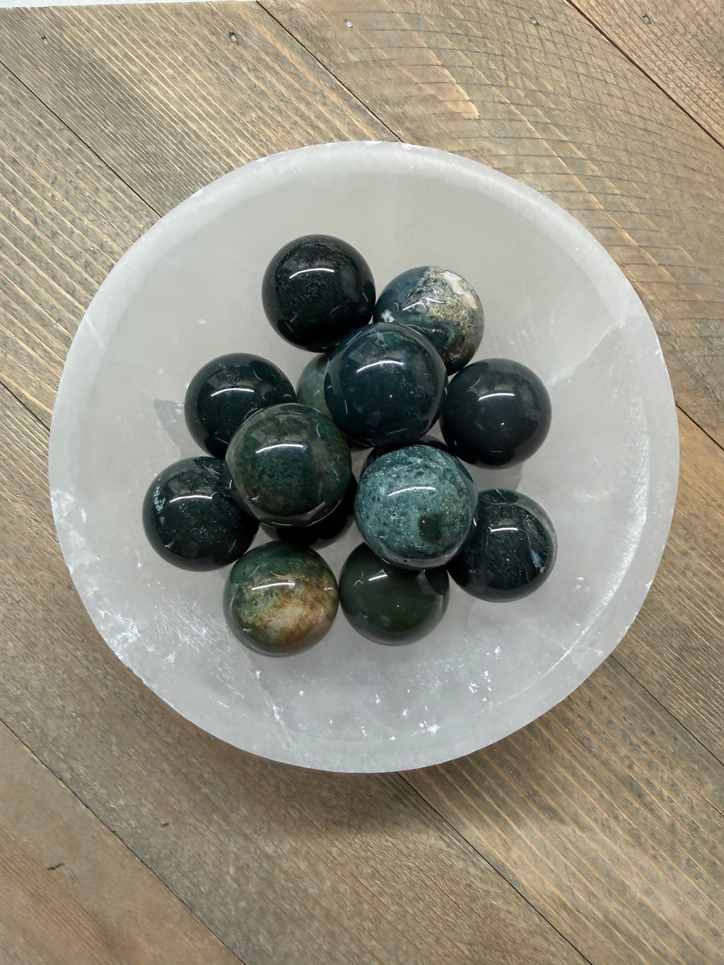 A group of polished dark green and black gemstone balls arranged in a white ceramic bowl on a rustic wooden surface.