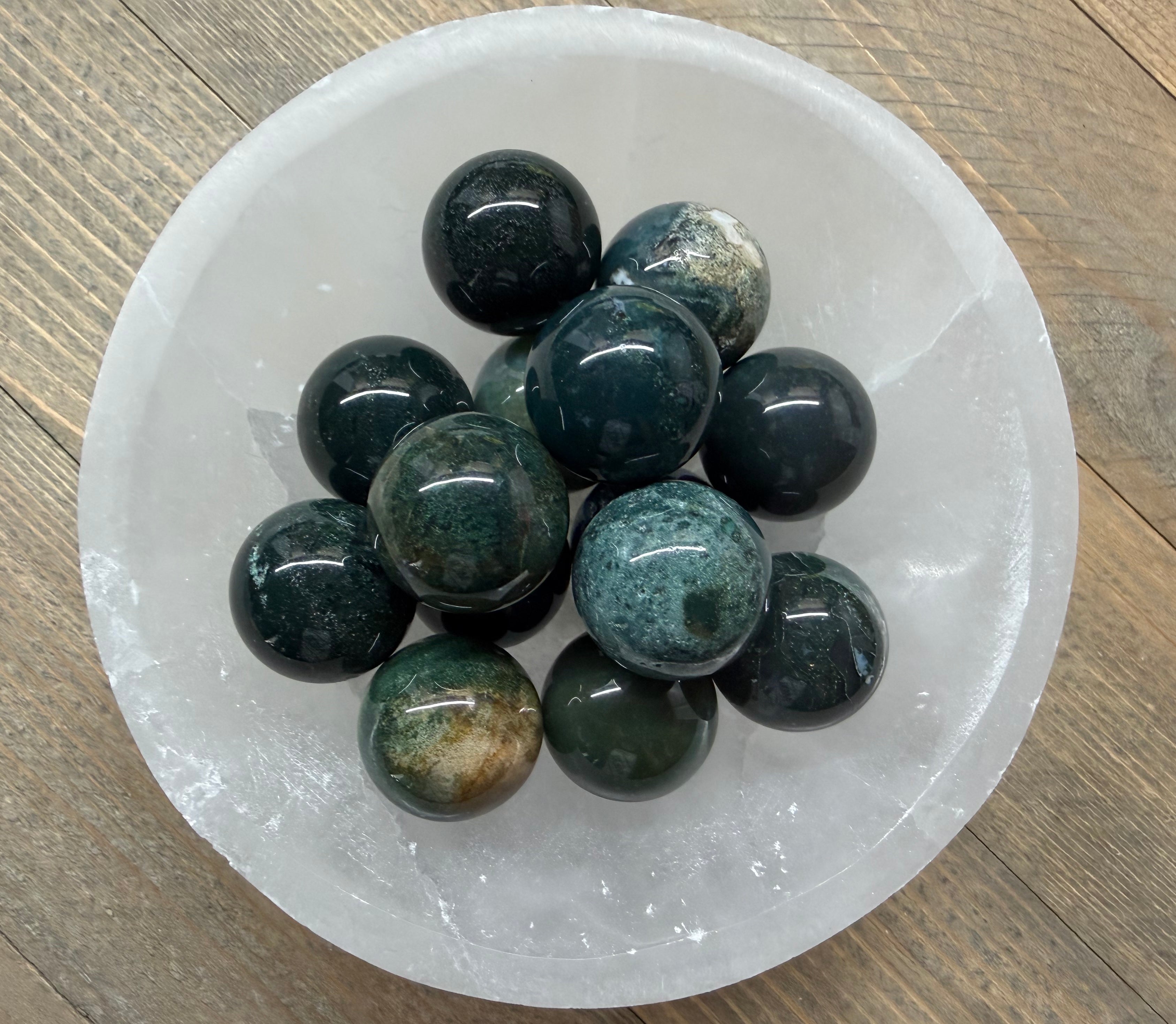 A cluster of polished dark green and black jasper gemstone balls arranged inside a white crystal bowl on a light wood surface.