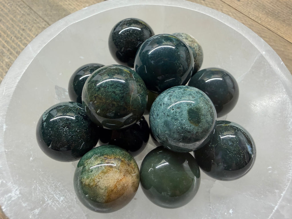 A group of polished dark green and black jasper stone balls with natural mottled patterns, arranged inside a clear glass bowl on a wooden surface.