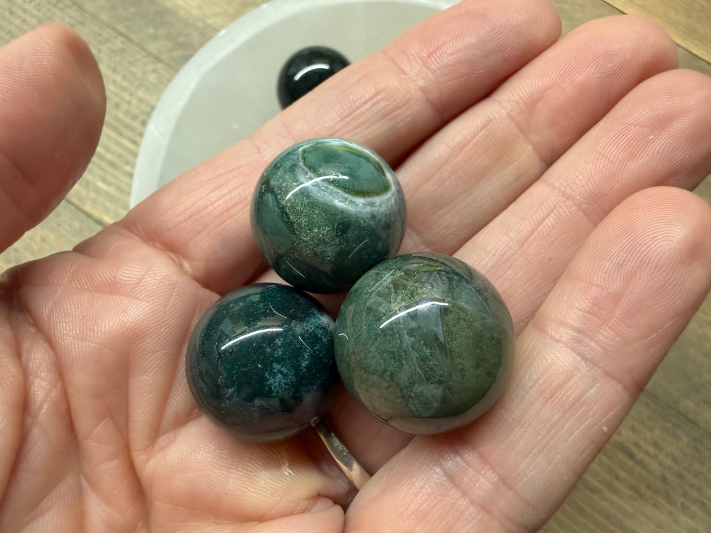 Three polished green stone beads held in a person's hand, with a white bowl and wood surface in the background.