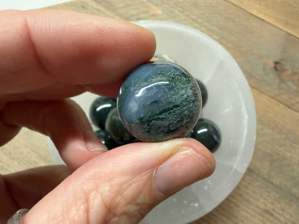 A hand holding a polished oval gemstone with a blue-gray and green marbled pattern, with more similar stones in a white dish on a wooden surface in the background.
