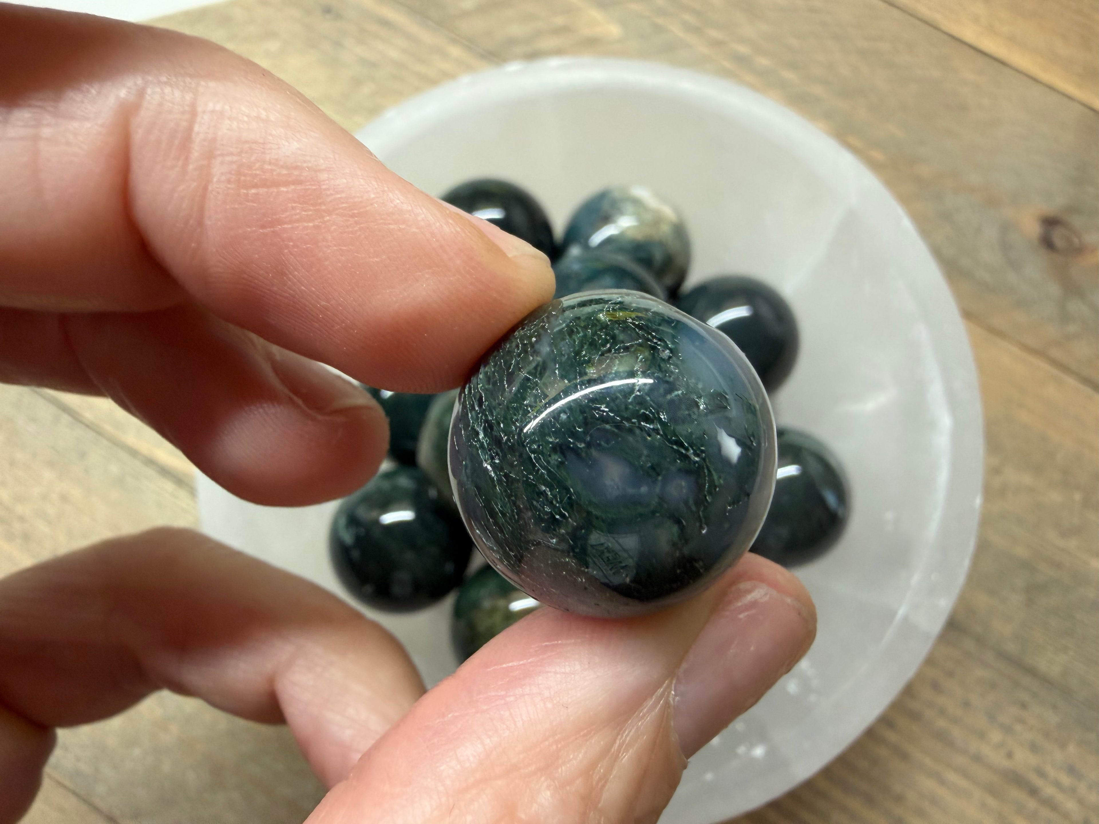 A hand holds a polished dark green stone sphere in front of a white bowl containing several similar stone balls on a wooden surface.