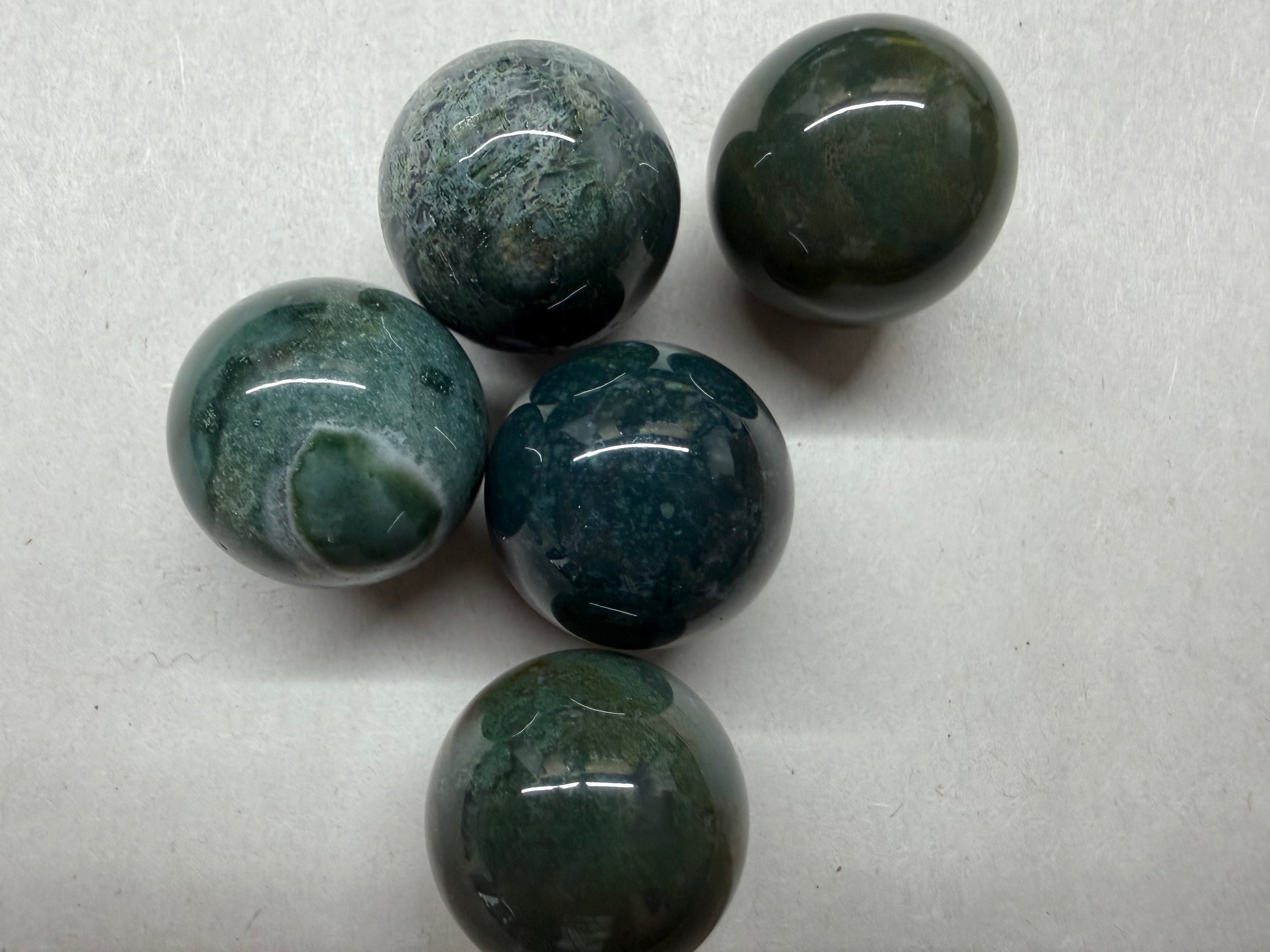 Five polished dark green and blue-green stone spheres with natural marbled patterns, arranged on a light gray textured background.