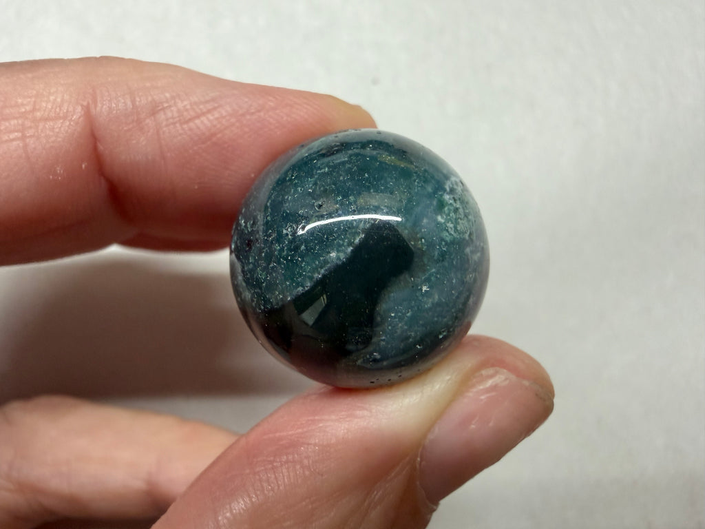 A hand holding a polished round gemstone with a deep blue-green color and white speckled inclusions, set against a plain light background.