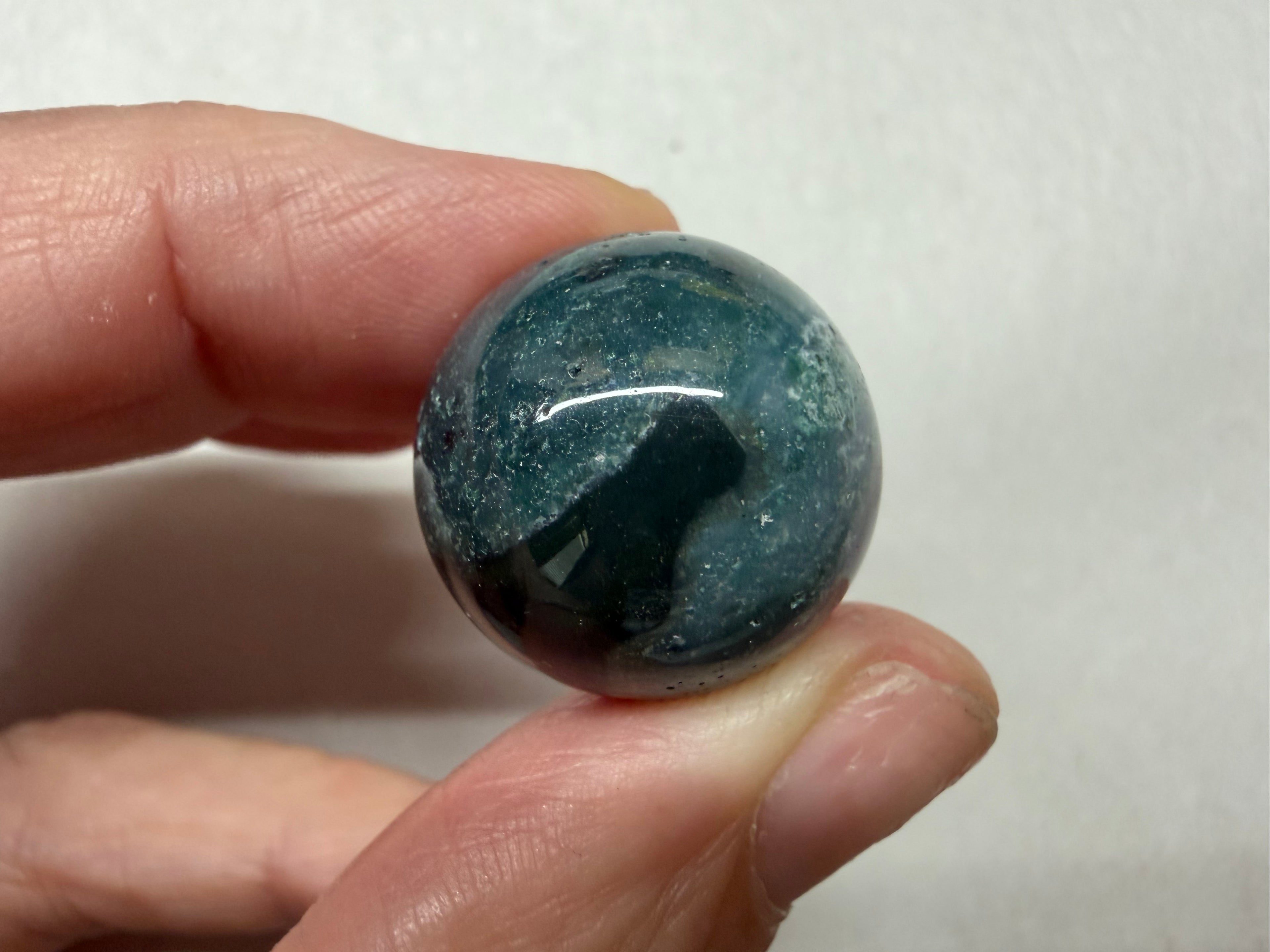 A hand holding a polished round gemstone with a deep blue-green color and white speckled inclusions, set against a plain light background.