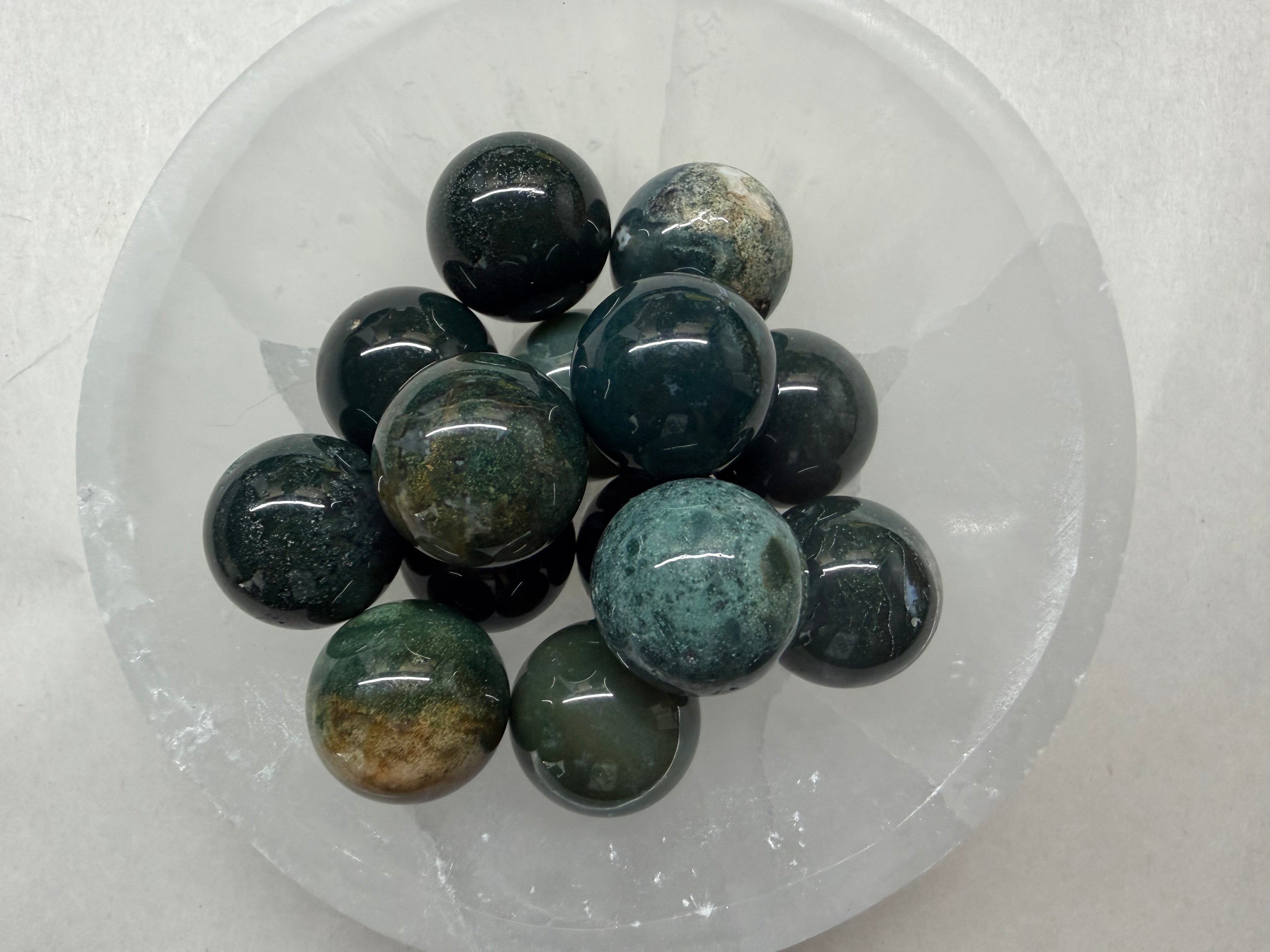 A cluster of polished dark green and black stone balls with natural mottled patterns, displayed inside a clear, round plastic container on a white textured background.