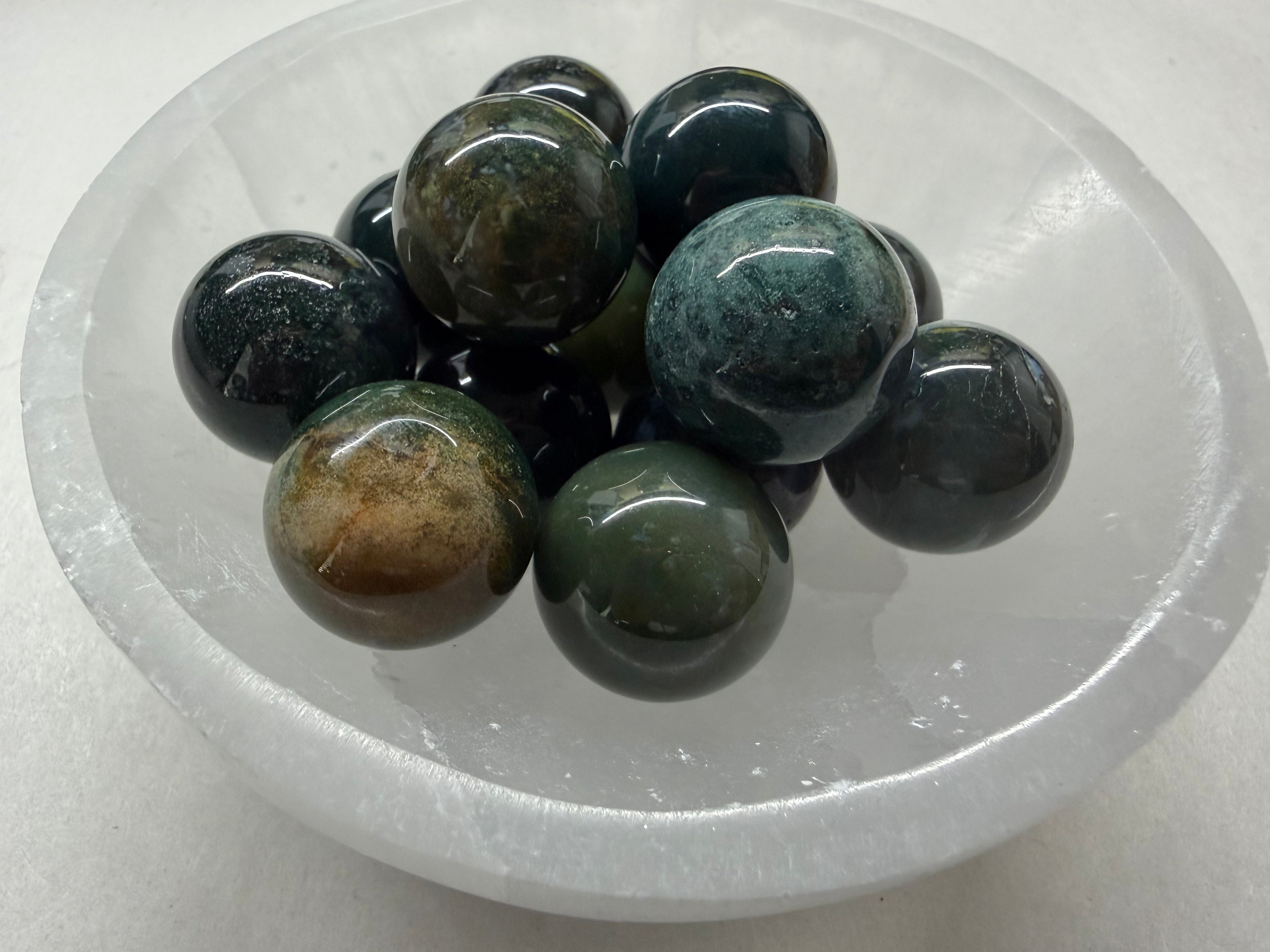 A cluster of polished dark green and black stone beads with natural inclusions, displayed inside a clear, oval-shaped glass bowl on a light gray background.