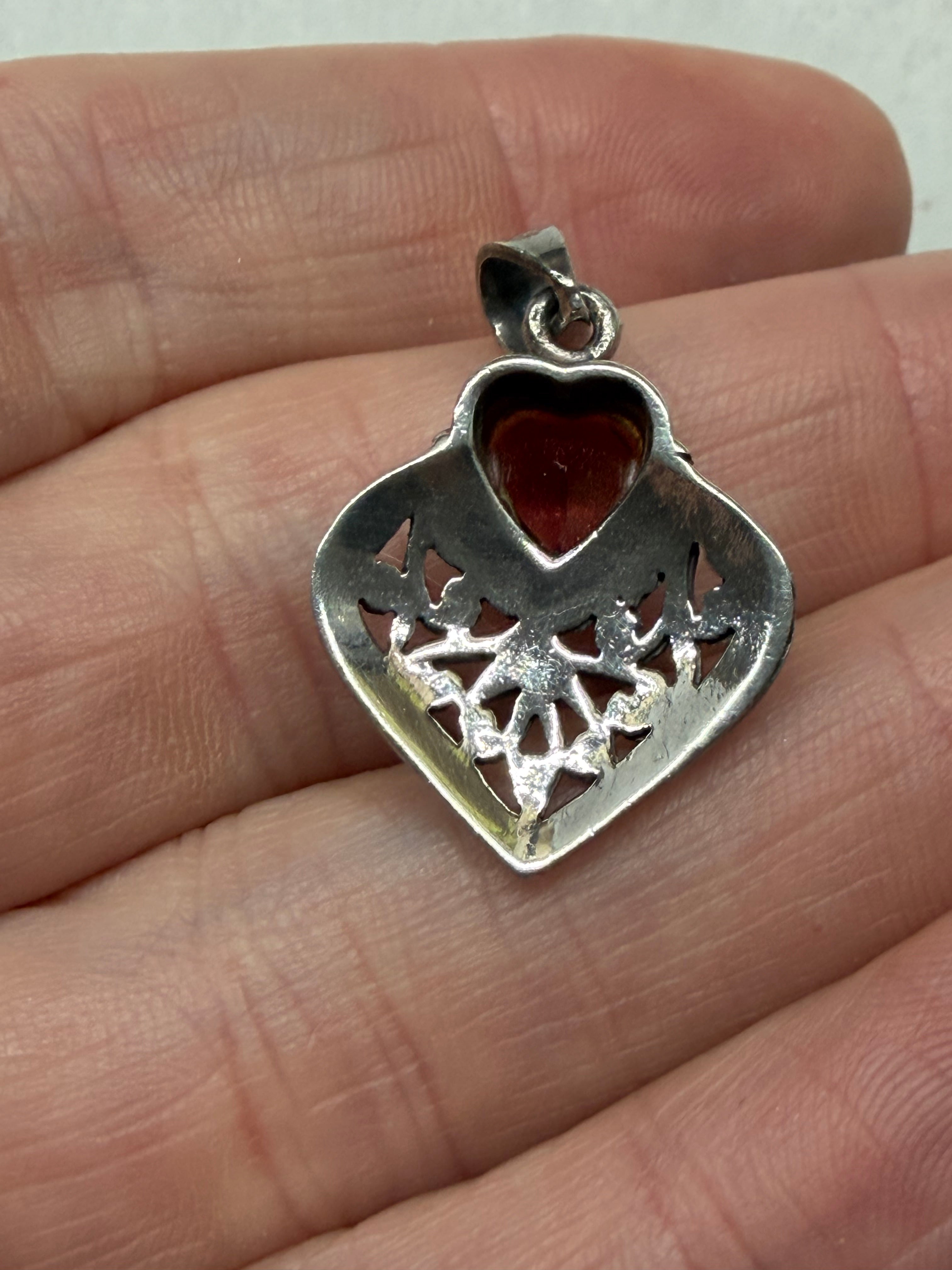 A silver heart-shaped pendant with a red heart-shaped stone, resting on a person's fingers to show its size.