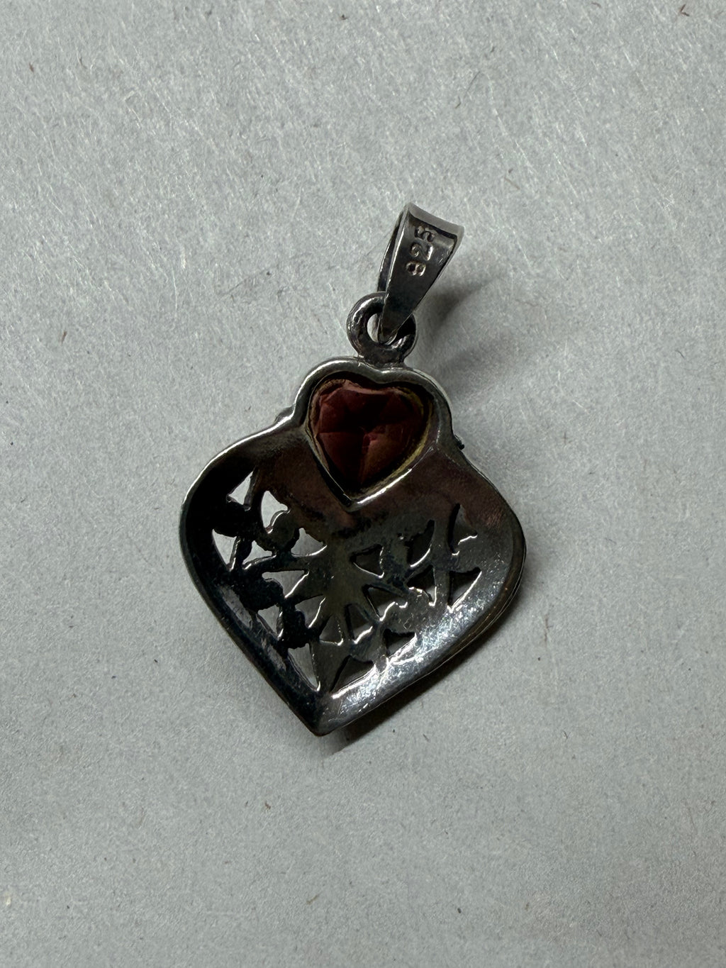 A heart-shaped filigree pendant in dark silver-tone metal with a small reddish-brown heart-shaped gemstone, shown on a textured light gray background.