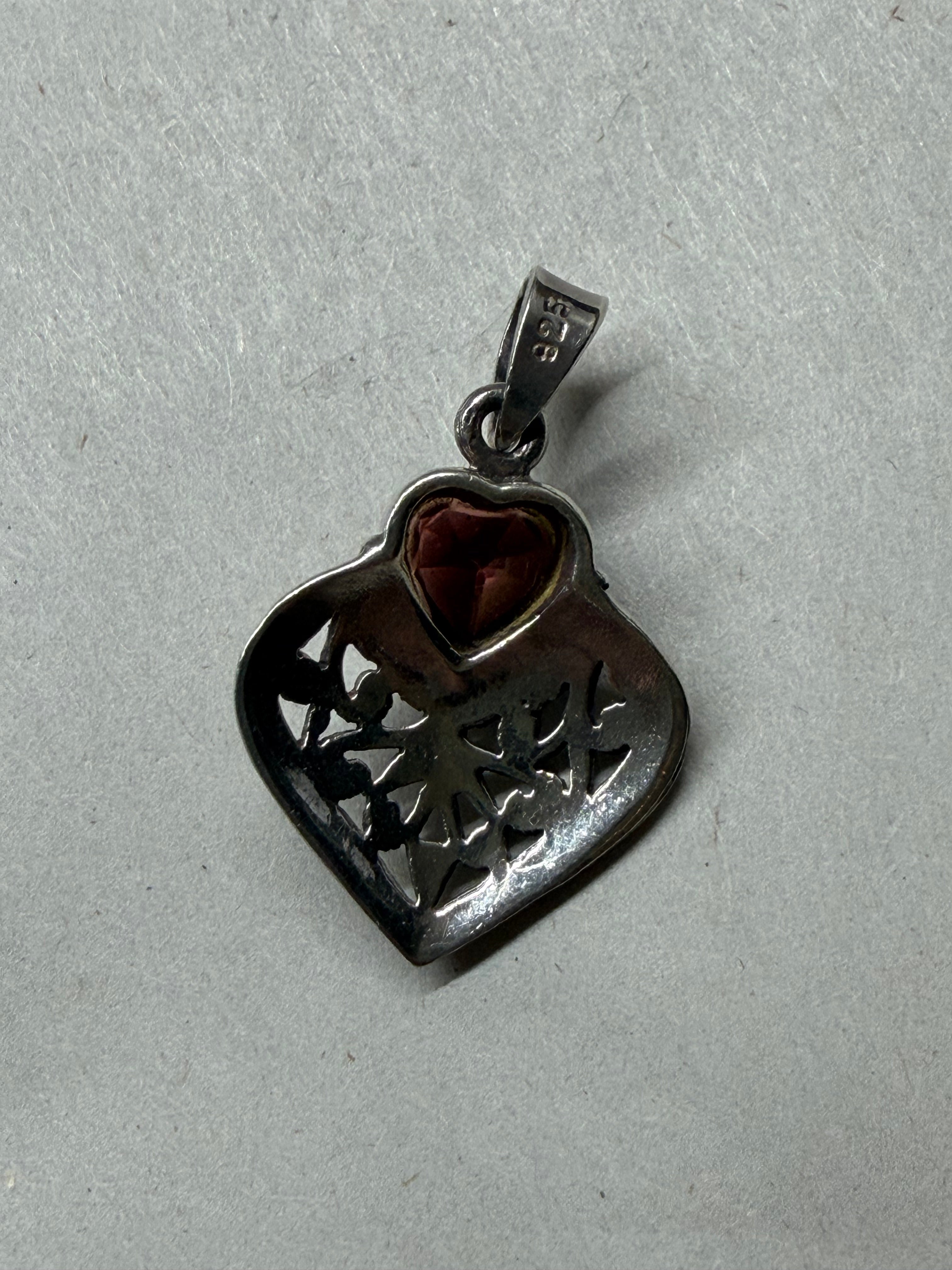 A heart-shaped filigree pendant in dark silver-tone metal with a small reddish-brown heart-shaped gemstone, shown on a textured light gray background.