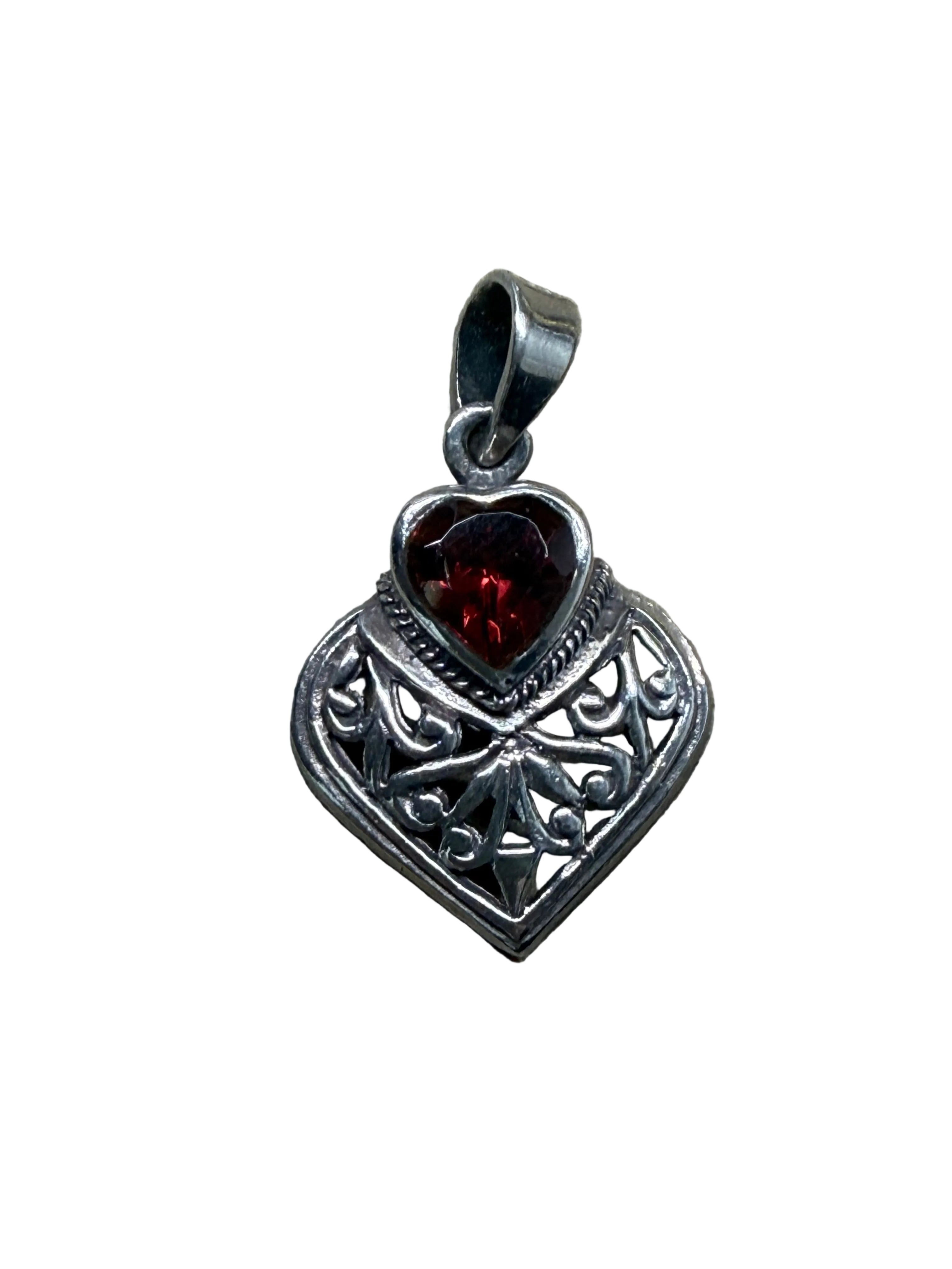 A heart-shaped silver pendant with an ornate filigree design and a dark red heart-shaped gemstone, resting on a rustic wooden surface.