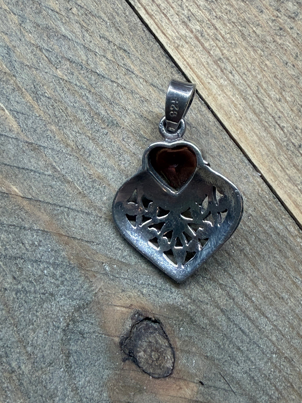 A silver heart-shaped pendant with a tree of life design and a small red heart inlay, resting on a weathered wooden surface.