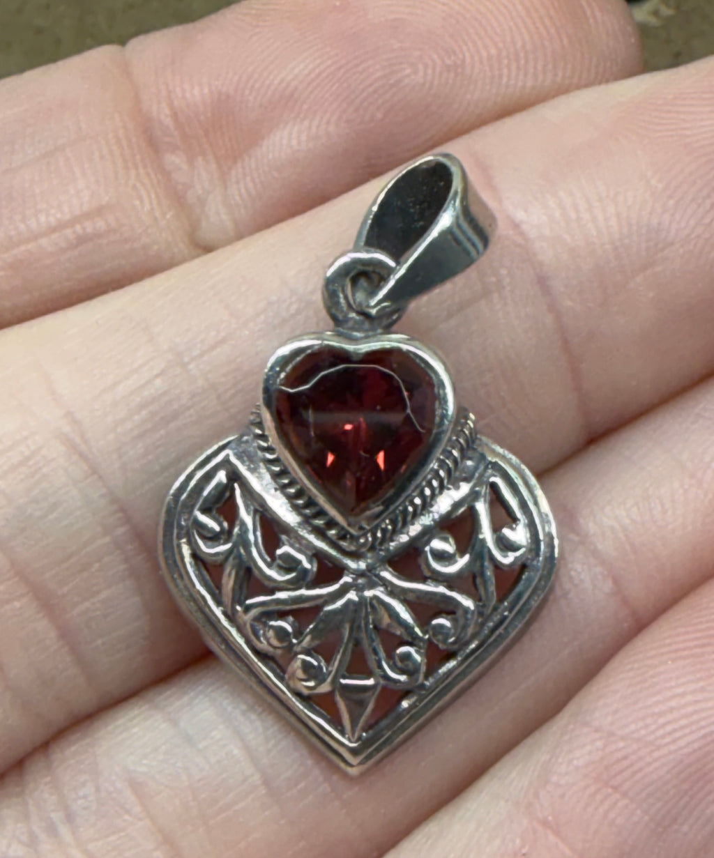 A silver filigree heart pendant with a faceted dark red heart-shaped gemstone, shown resting on a person's fingers.