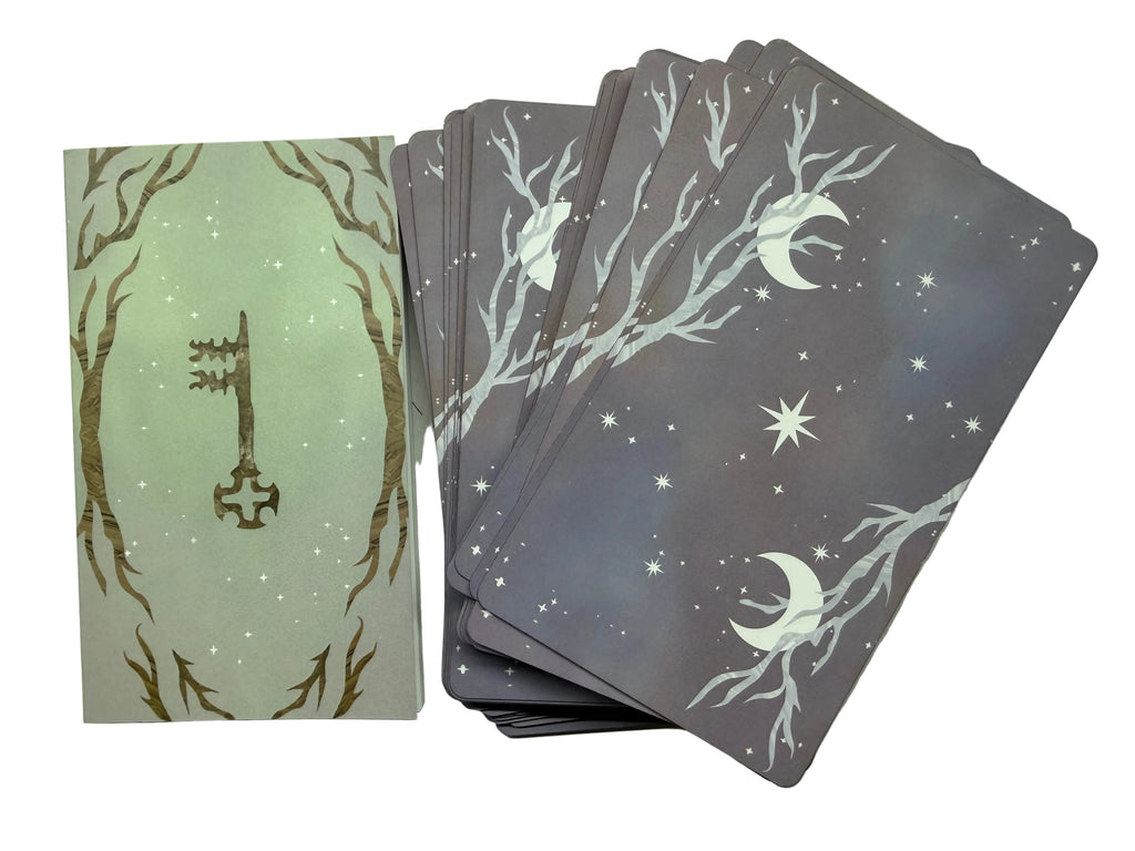 A tarot card deck with a light sage green front card featuring a gold key, surrounded by bare branches, shown next to a fanned stack of back cards in a muted purple-gray color with white moon, star, and branch illustrations.