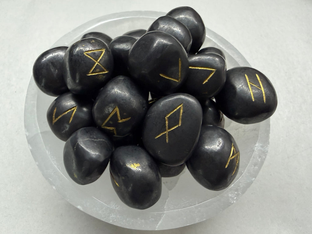 A pile of smooth black tumbled stones with engraved gold runic symbols, arranged in a clear glass bowl on a light gray surface.