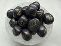 A pile of smooth black tumbled stones with engraved gold runic symbols, arranged in a clear glass bowl on a light gray surface.