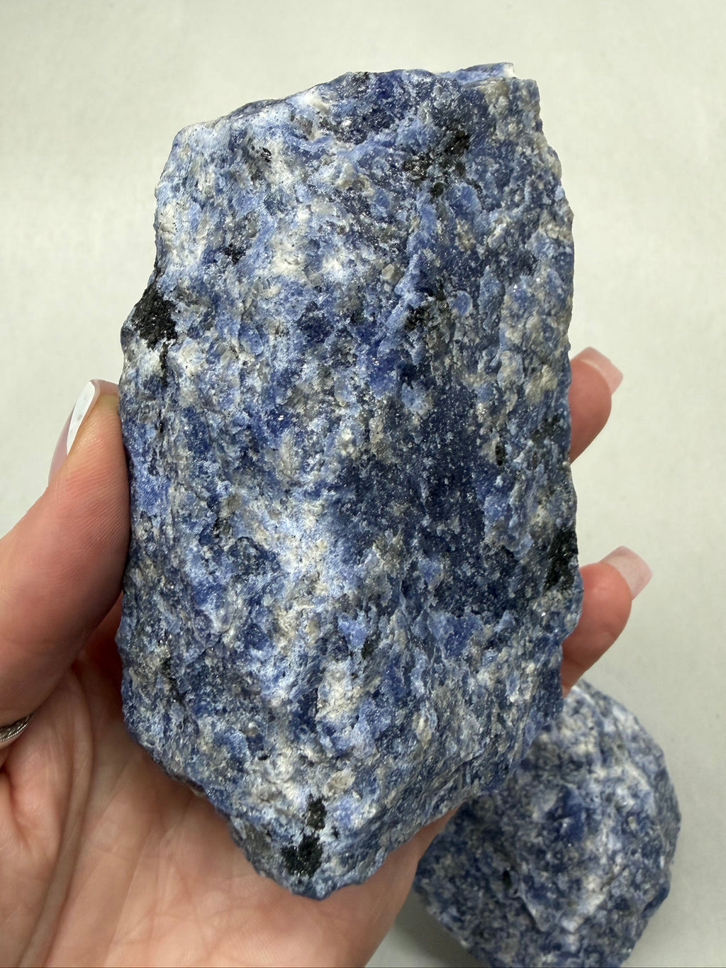 A hand holding a rough blue and white speckled stone or mineral specimen, with a second similar stone visible in the background against a plain light backdrop.