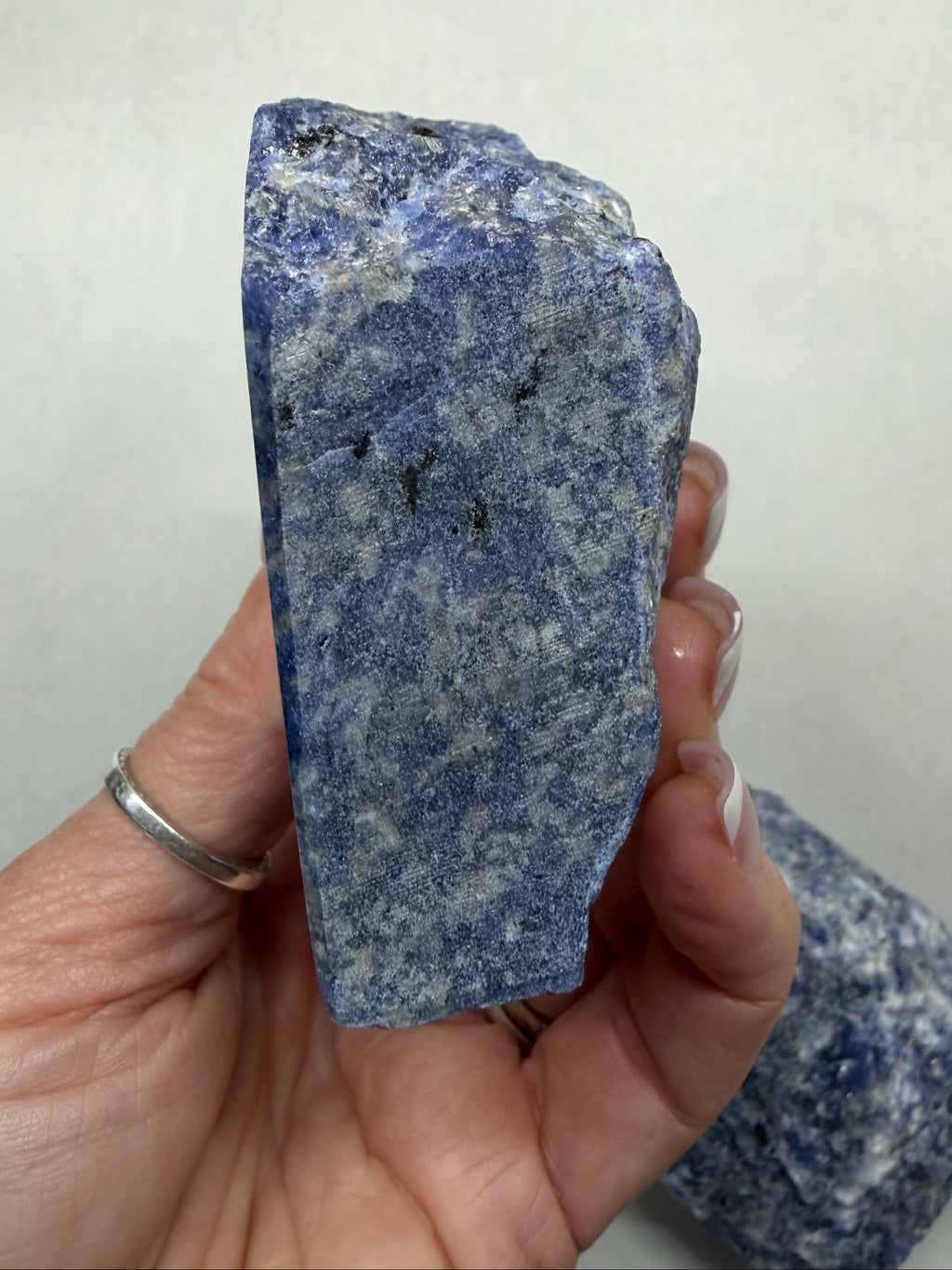 A hand holding a rough blue and white speckled stone, with a second similar stone visible in the background against a plain light backdrop.