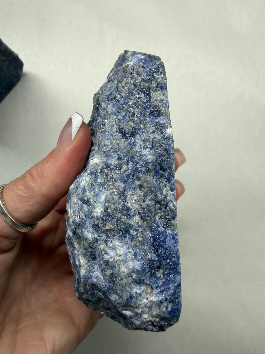 A hand holding a rough, unpolished blue and white speckled stone or mineral against a plain light background.
