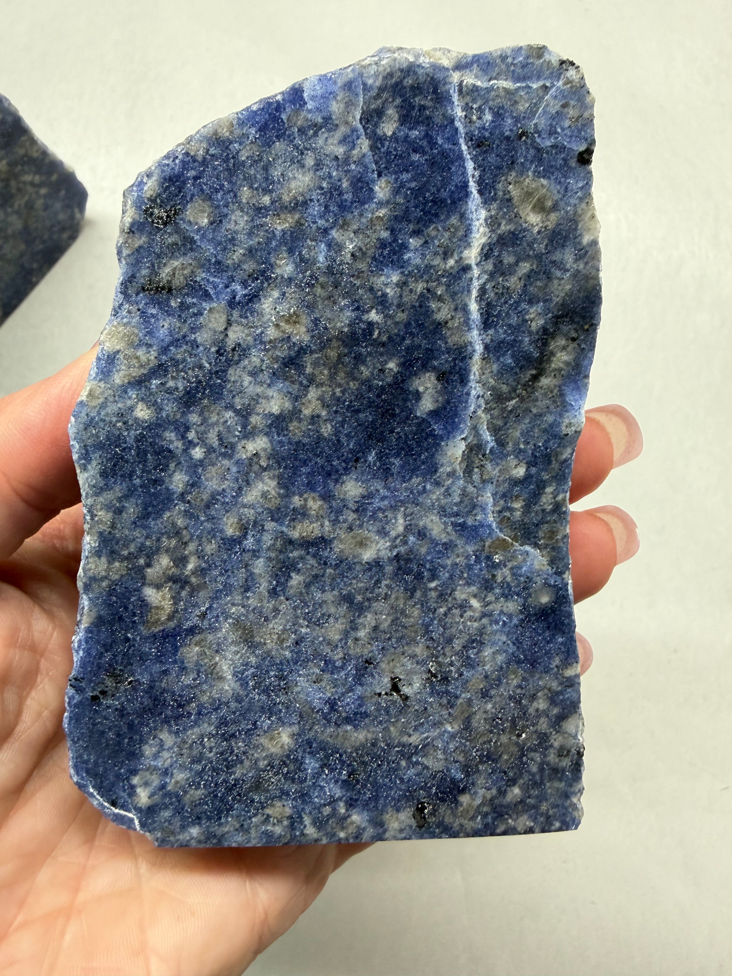 A hand holding a raw, rough-cut blue lapis lazuli crystal specimen with natural white and gray flecks, against a plain off-white background.