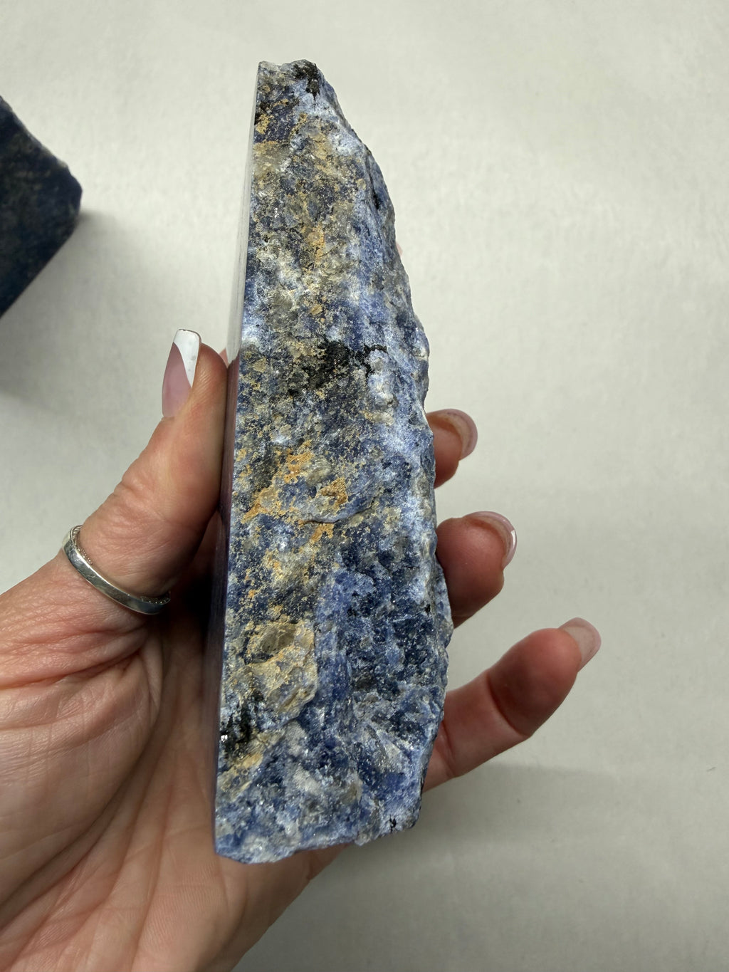 A hand holding a rough, raw mineral or crystal specimen with a speckled blue, gray, black, and tan texture against a plain off-white background.