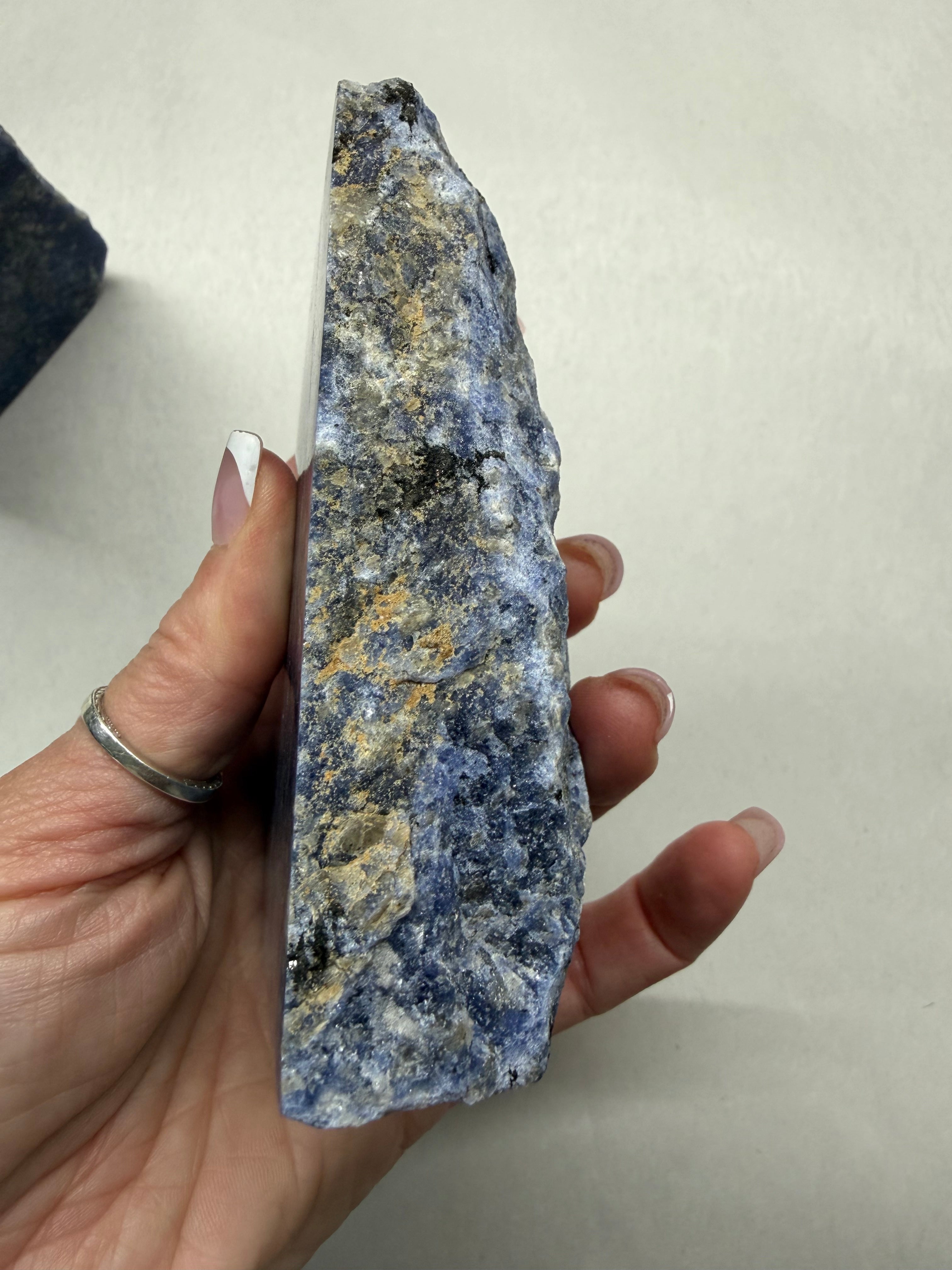 A hand holding a rough, raw mineral or crystal specimen with a speckled blue, gray, black, and tan texture against a plain off-white background.