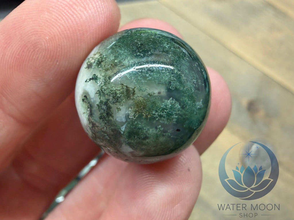Green marbled stone held between fingers with 'Water Moon Shop' logo in the corner.