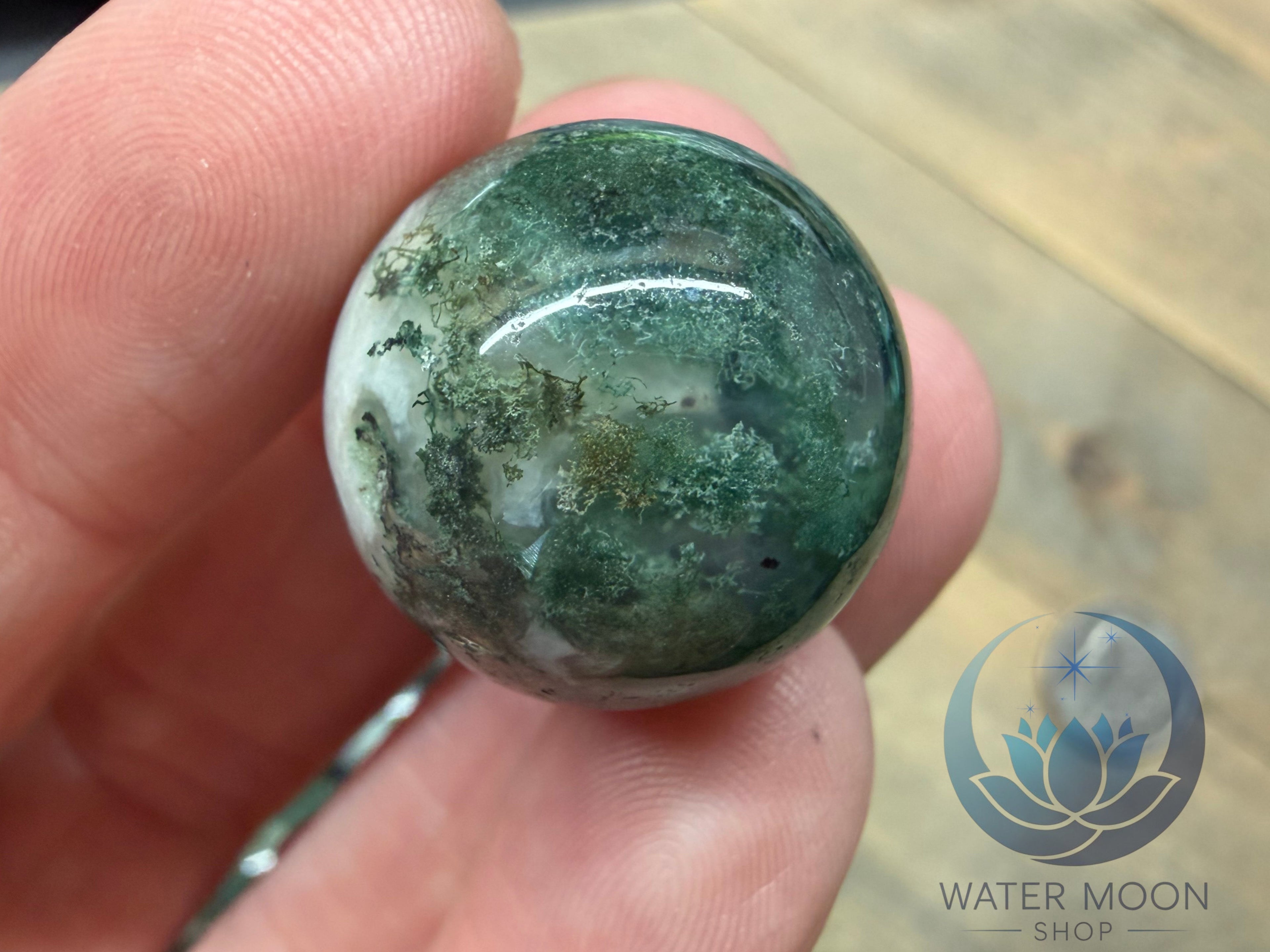 Green marbled stone held between fingers with 'Water Moon Shop' logo in the corner.